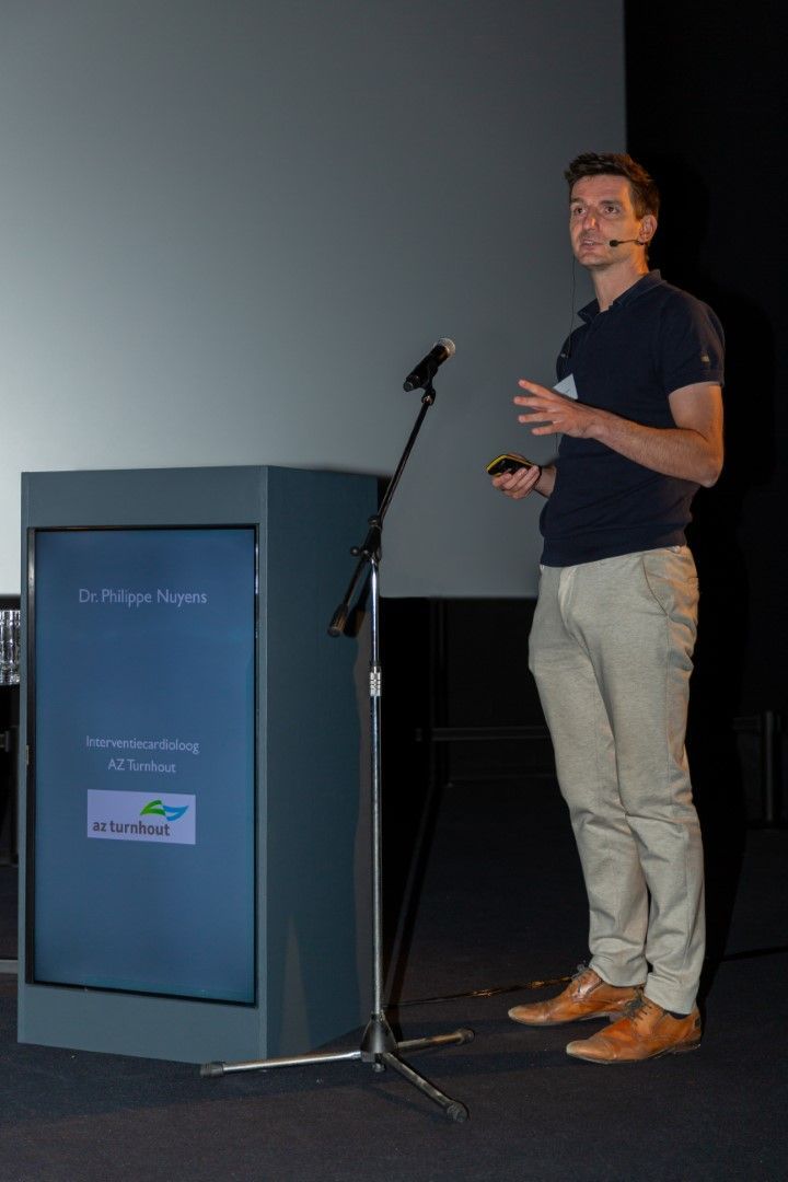 Een man spreekt op een podium naast een lessenaar en microfoon in een donkere zaal.