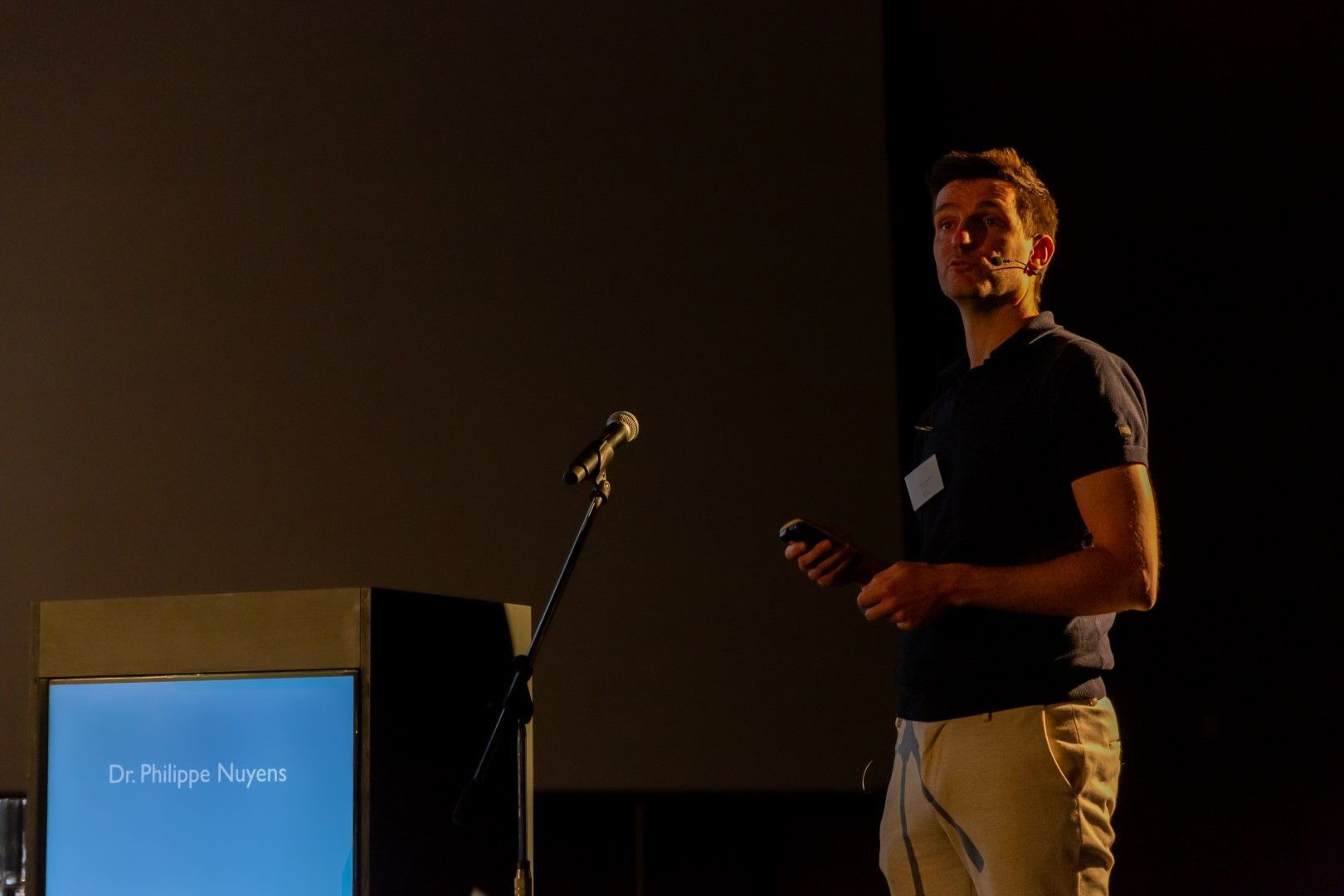 De presentator staat op het podium naast een microfoon en lessenaar, met een dia die op een donker scherm wordt geprojecteerd.