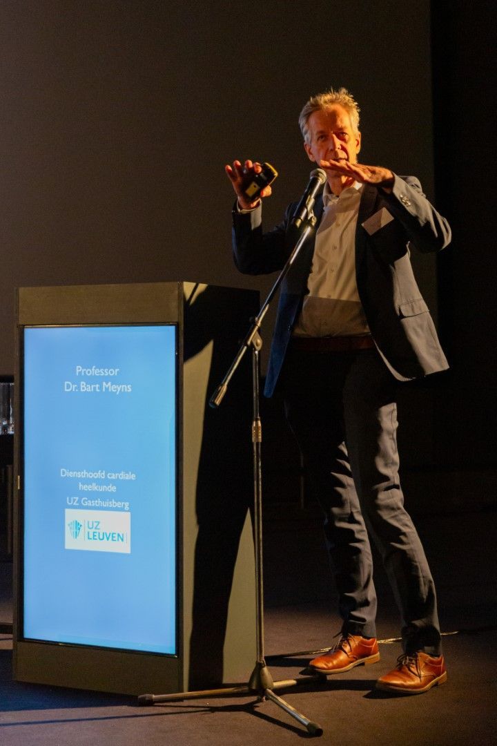 Spreker achter een podium op een donker toneel, gebarend naast een blauw presentatiescherm.