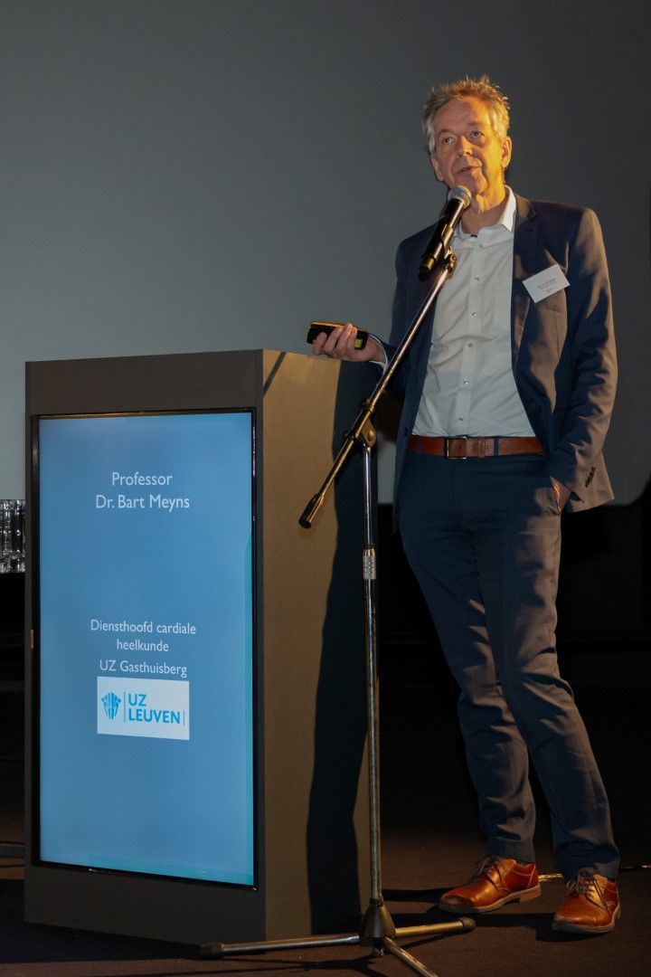 Een spreker staat achter een lessenaar op een podium en geeft een presentatie bij gedempt licht.