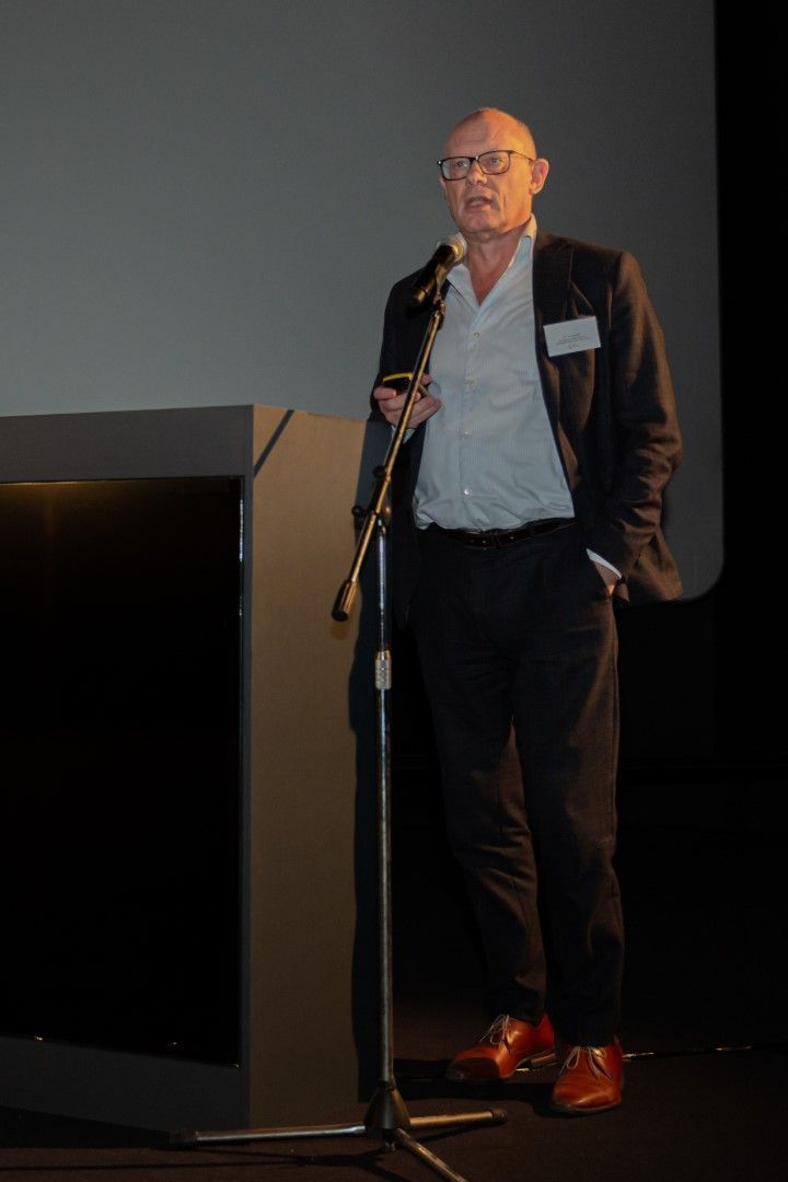 Een man spreekt vanaf een podium op een donker toneel, met een wandelstok in de hand en gekleed in een pak.