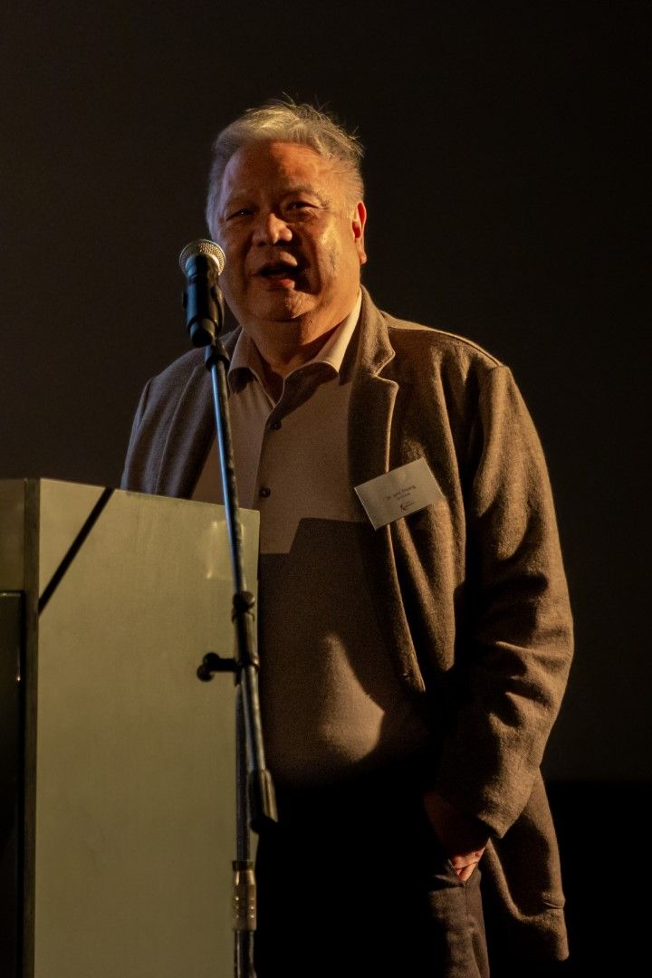 Een man spreekt achter een podium met een microfoon op een schemerig verlicht toneel, gekleed in een jas en een trui.