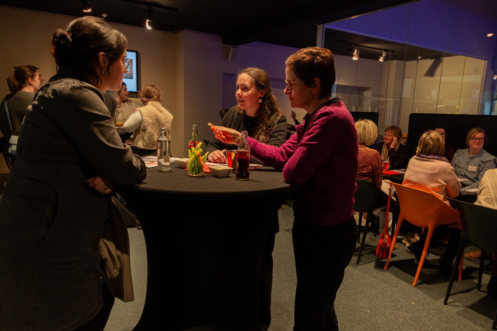 Mensen praten met elkaar rond een hoge ronde tafel in een schemerig verlichte evenementenruimte binnen.