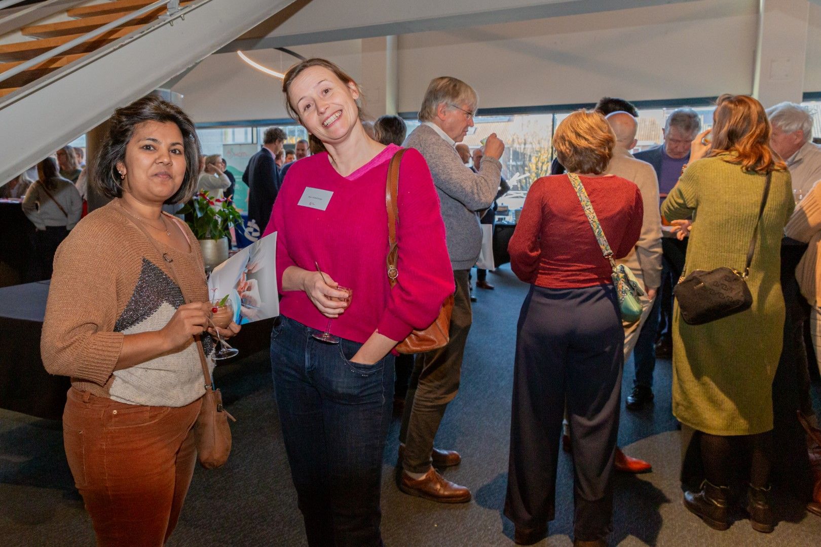 Mensen staan te praten in een witte evenemententent, met twee vrouwen op de voorgrond die papieren vasthouden.