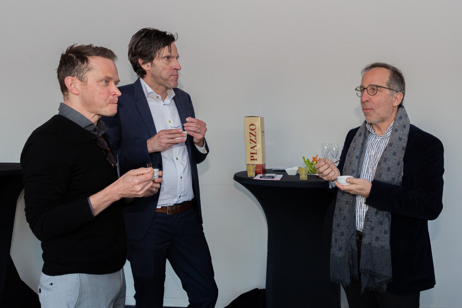 Drie personen in zakelijke kleding staan te praten aan een staande tafel tijdens een netwerkevenement.