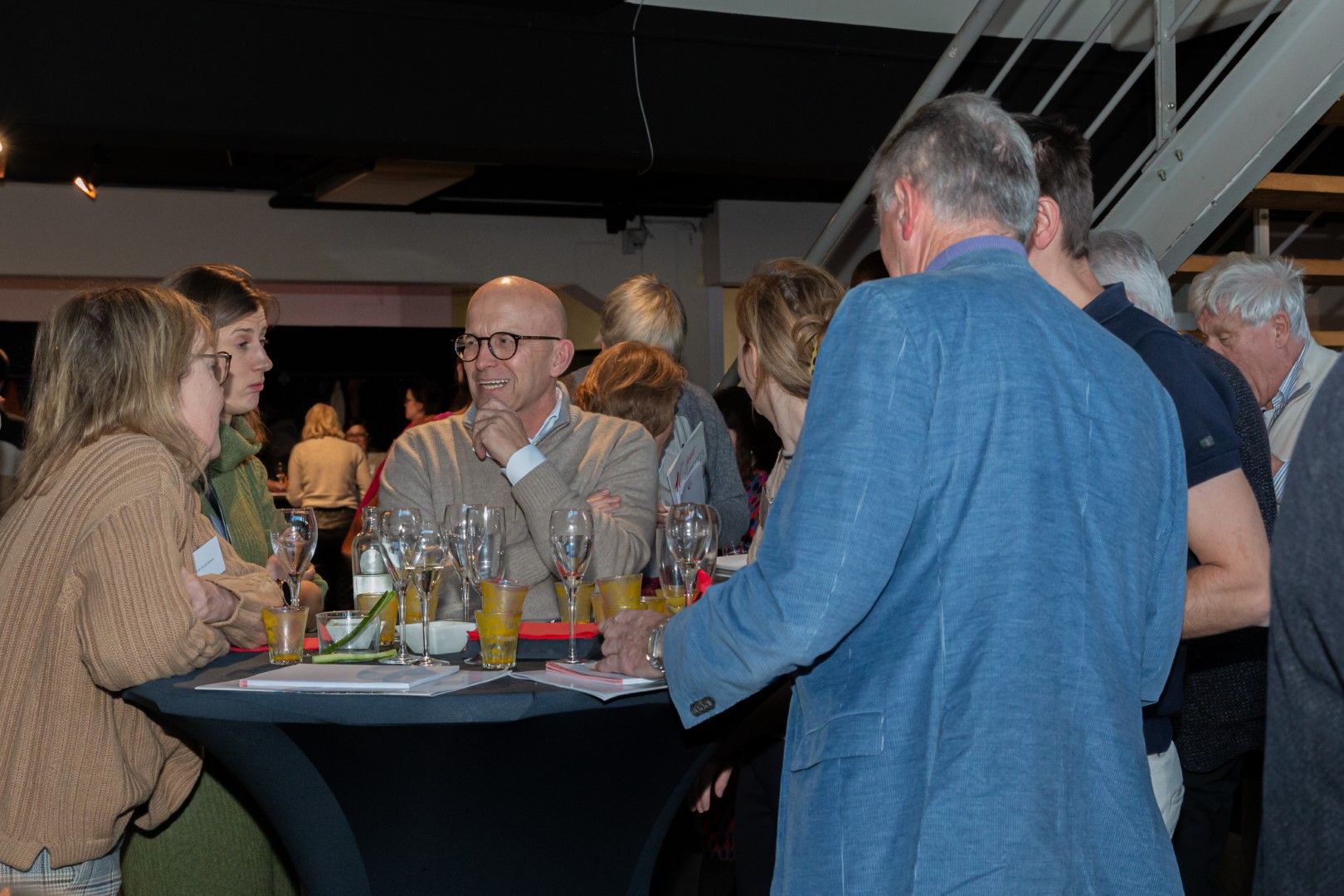 Mensen die gezellig met elkaar praten aan een cocktailtafel in een schemerige, overdekte evenementenruimte.