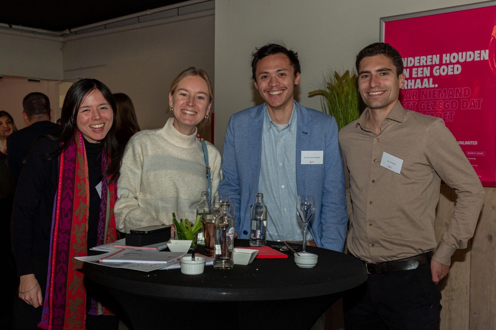Vier mensen glimlachen aan een kleine ronde tafel tijdens een evenement binnenshuis, met drankjes en een rode banner achter hen.