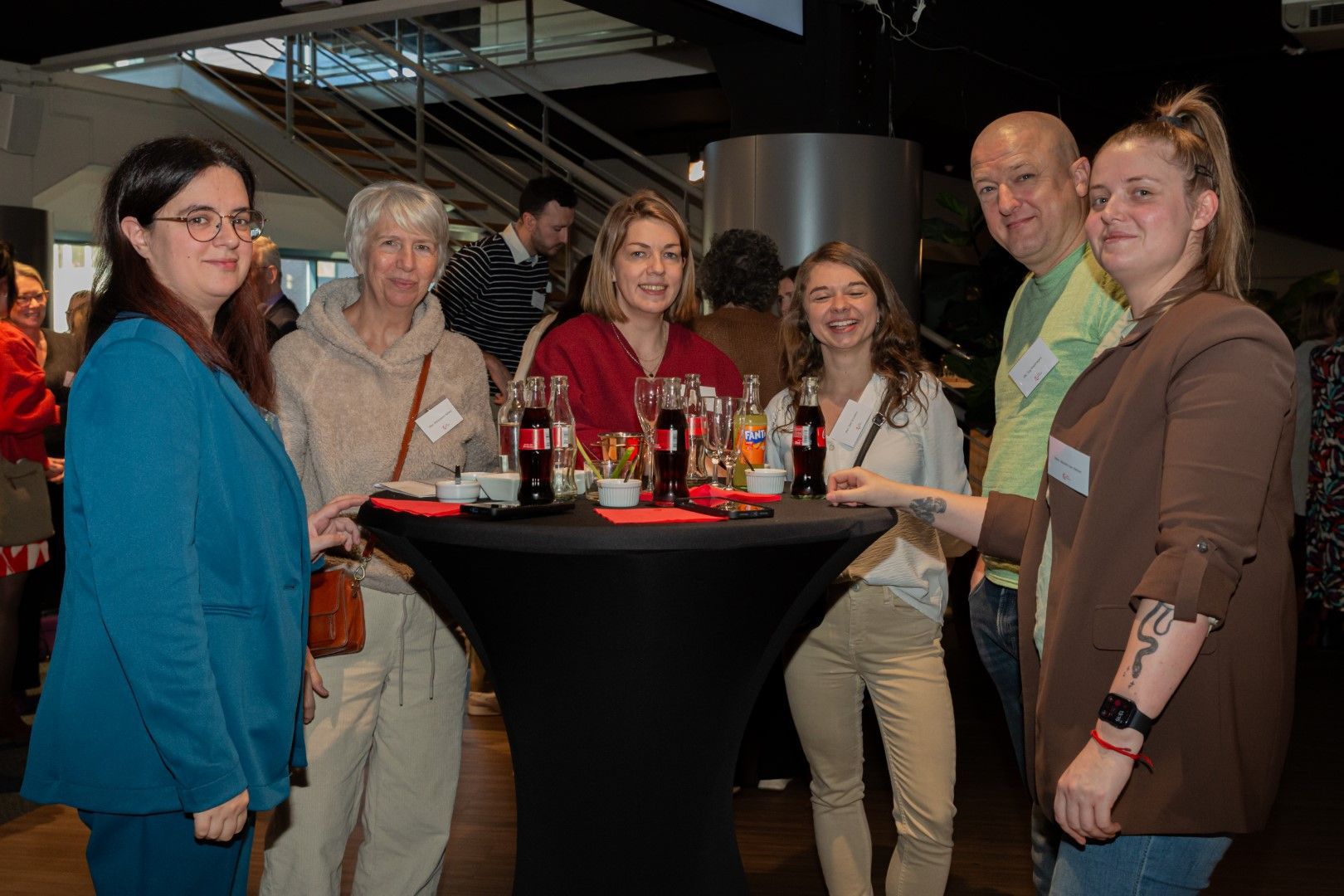 Zes mensen lachend rond een cocktailtafel met flessen en glazen tijdens een evenement binnenshuis.