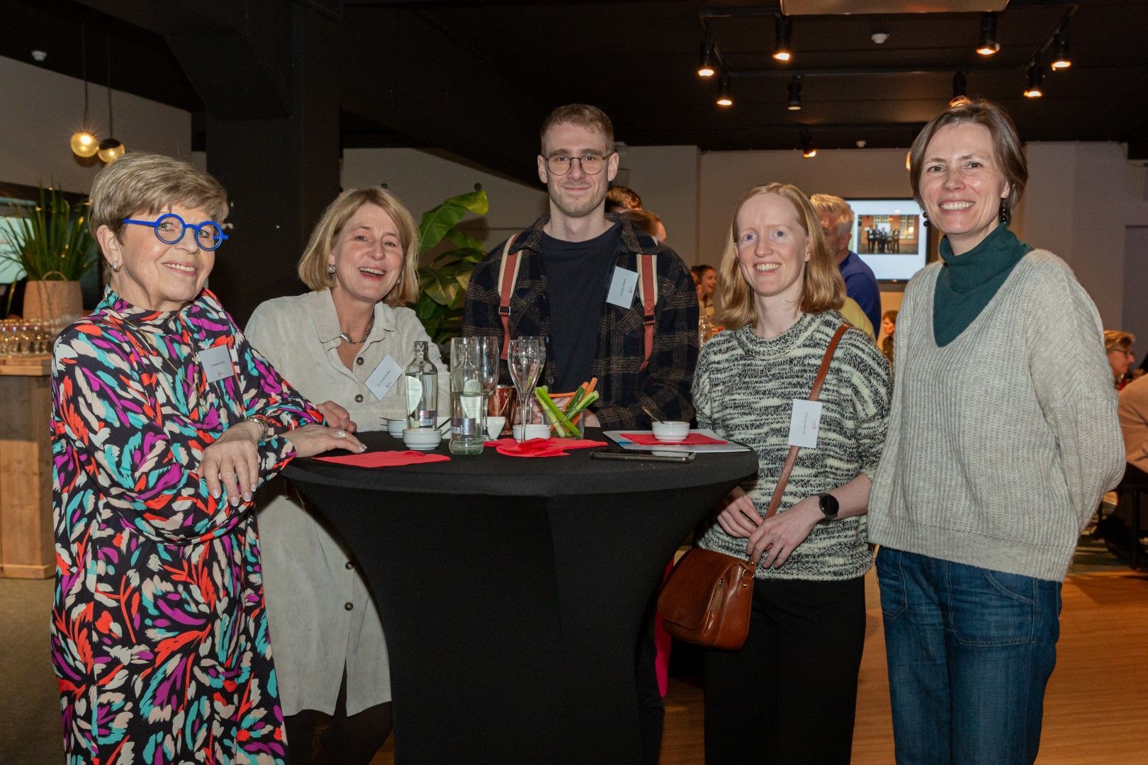 Vijf mensen staan rond een cocktailtafel tijdens een sociaal evenement binnenshuis en lachen naar de camera.