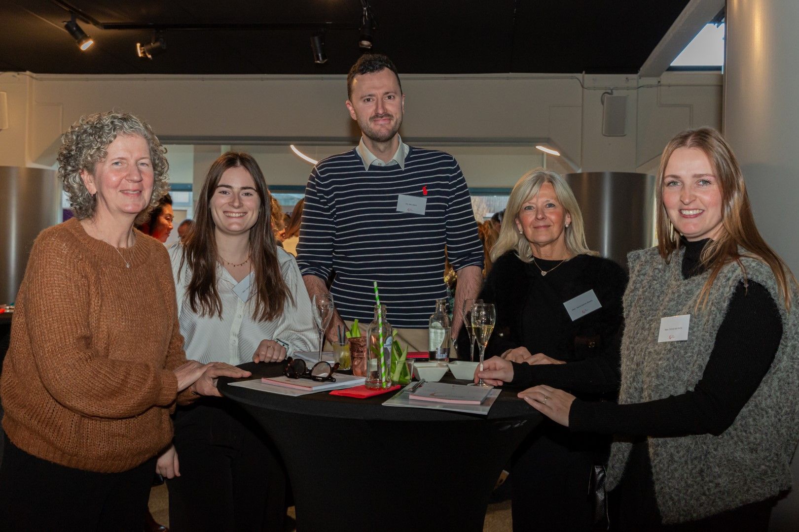 Vijf mensen zitten lachend rond een tafel bij een evenement binnenshuis, met drankjes en naambadges.