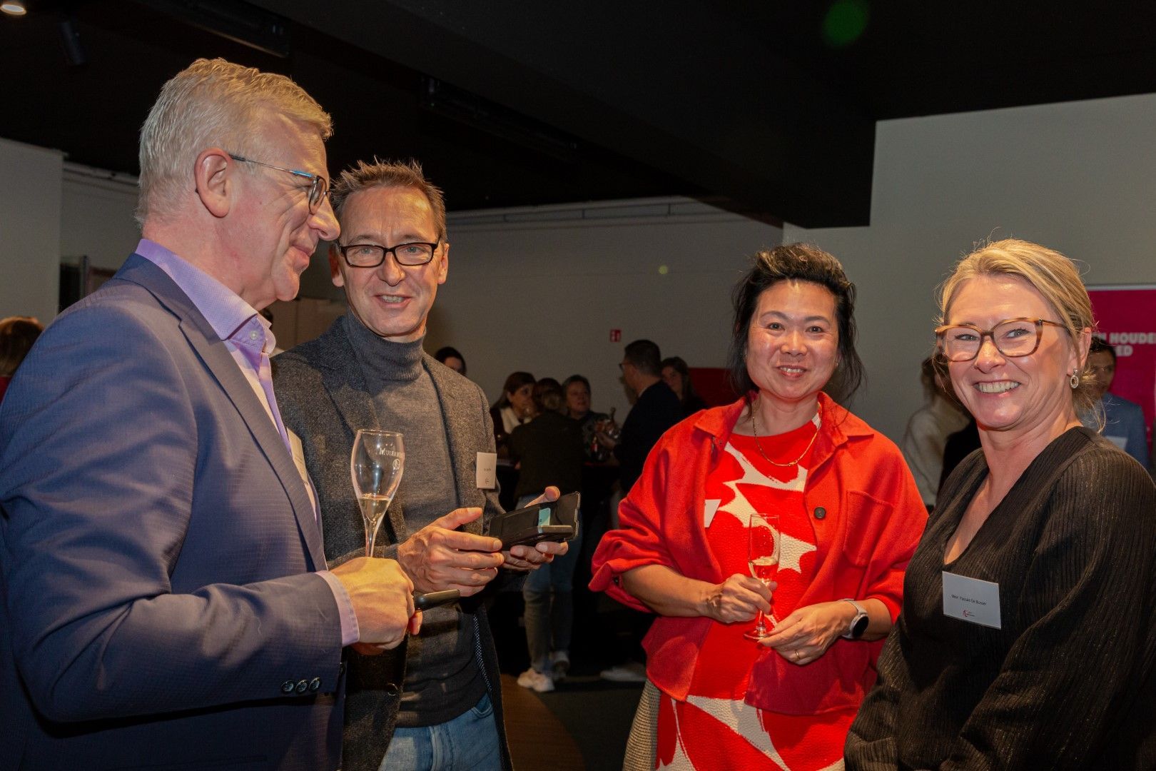 Vier mensen glimlachen en houden een drankje vast tijdens een gezellig samenzijn binnen.