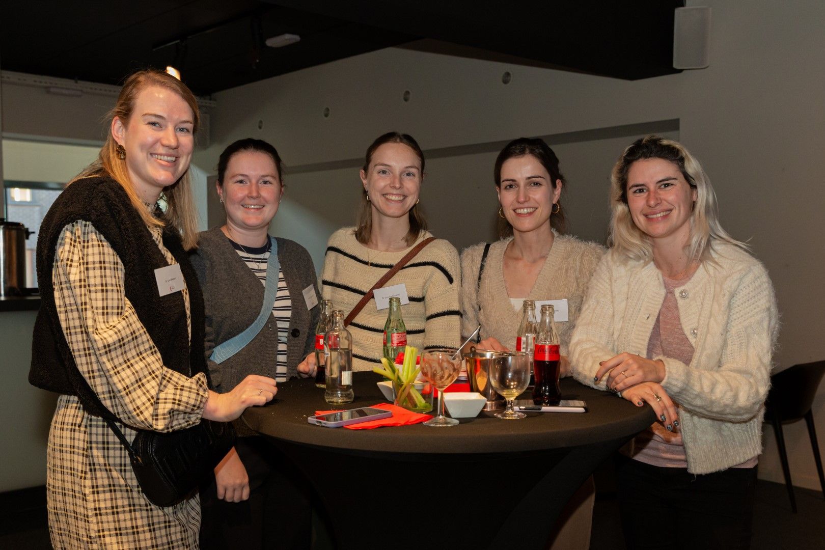 Vijf mensen zitten lachend rond een tafel met drankjes en eten in een schemerig verlicht restaurant.