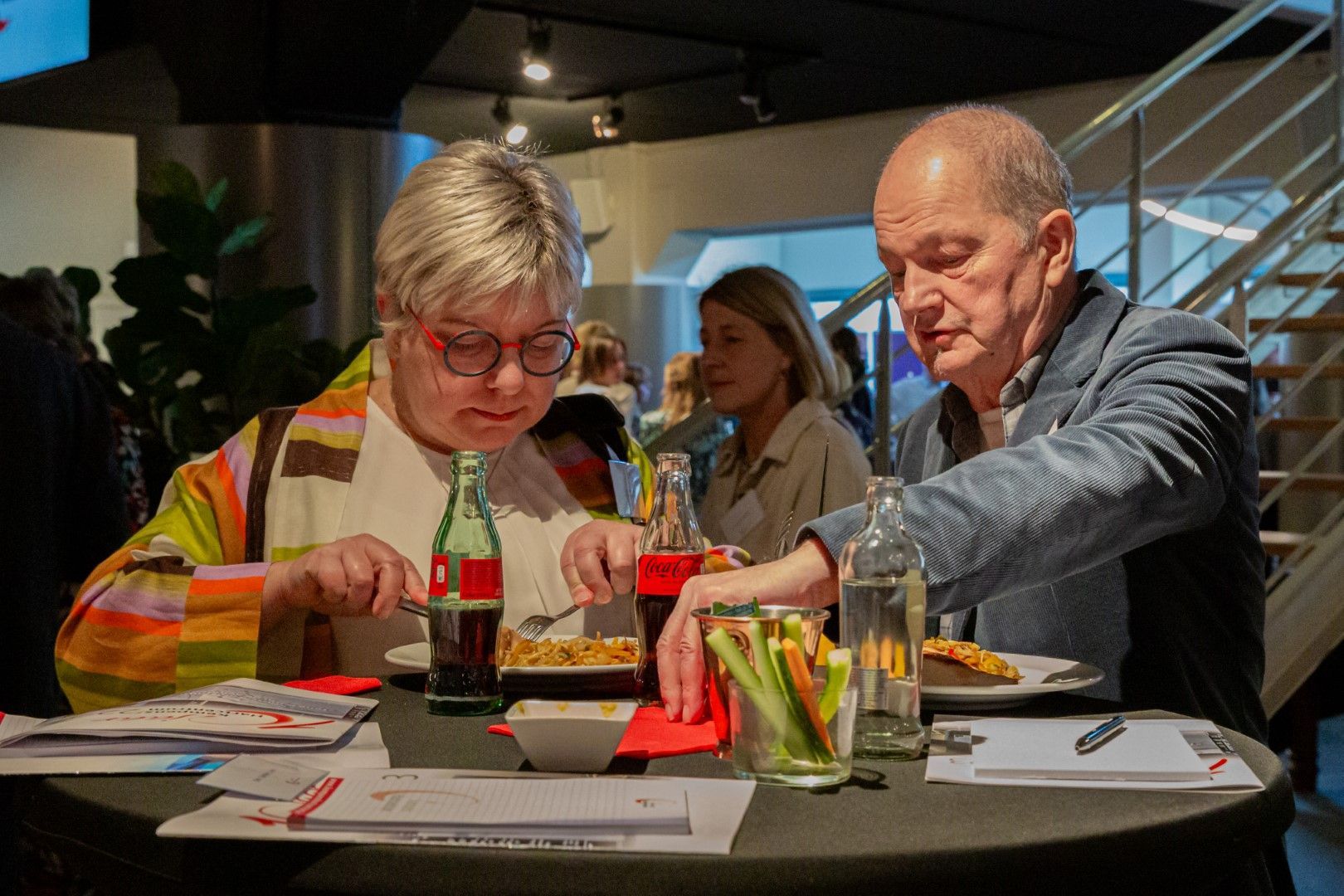 Twee mensen zitten aan een tafel en schilderen met felle kleuren, geconcentreerd bezig met een kunstactiviteit in een schemerig verlichte ruimte.