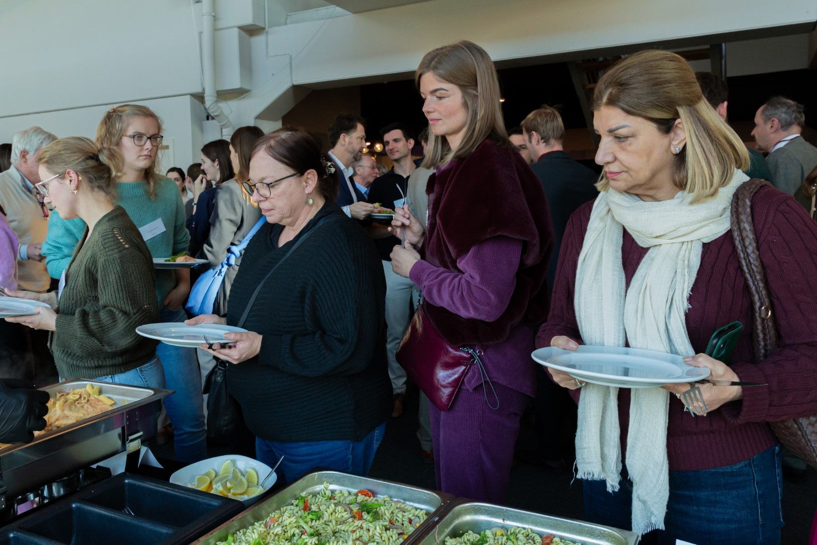 Mensen die eten serveren bij een buffet, met borden in hun handen, tijdens een druk evenement binnenshuis.
