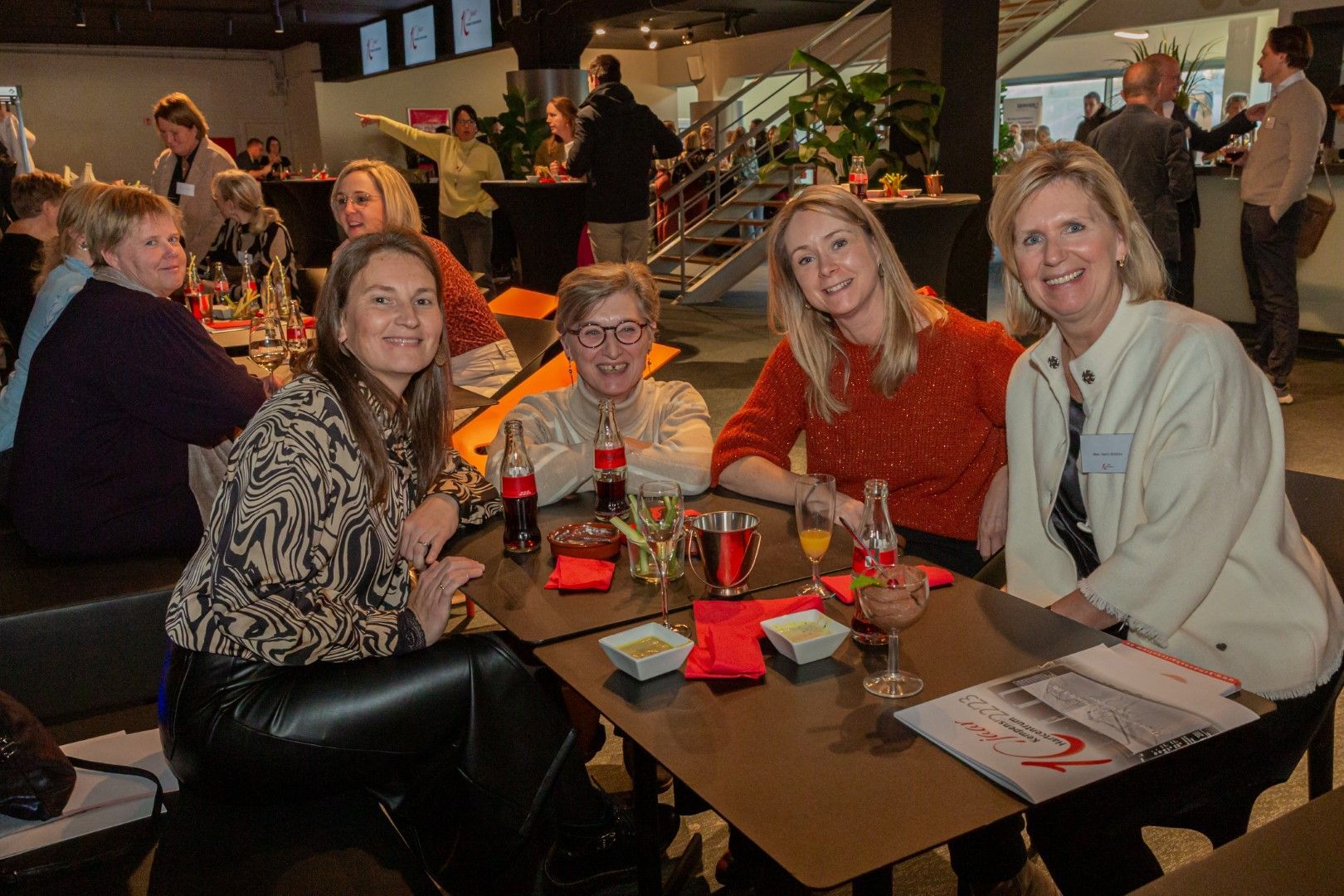 Vier mensen zitten aan een tafel in een schemerig restaurant, lachend en poserend met drankjes en menukaarten.