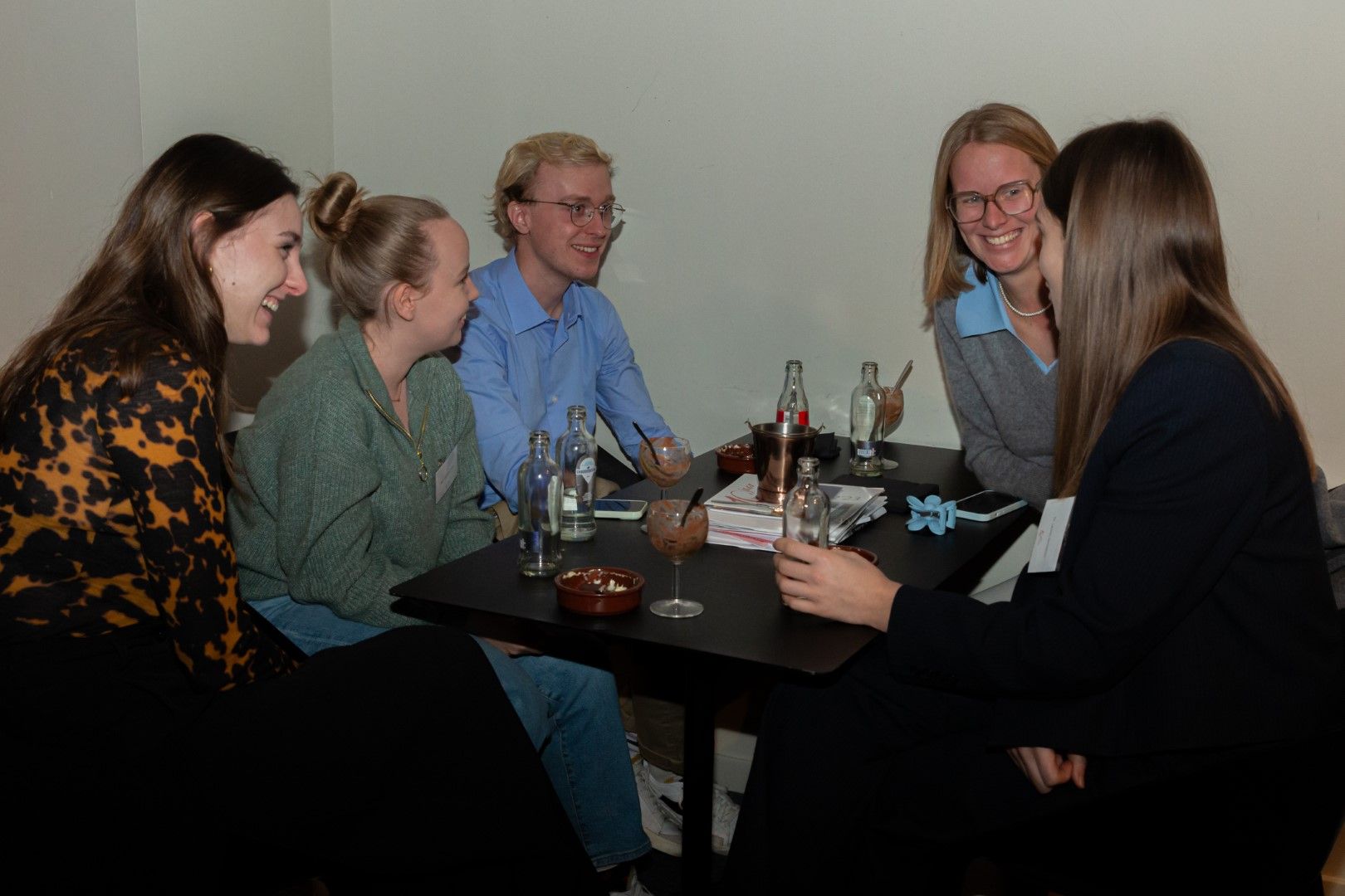 Vijf mensen die gezellig met elkaar praten rond een kleine tafel met een drankje in een informele binnenomgeving.
