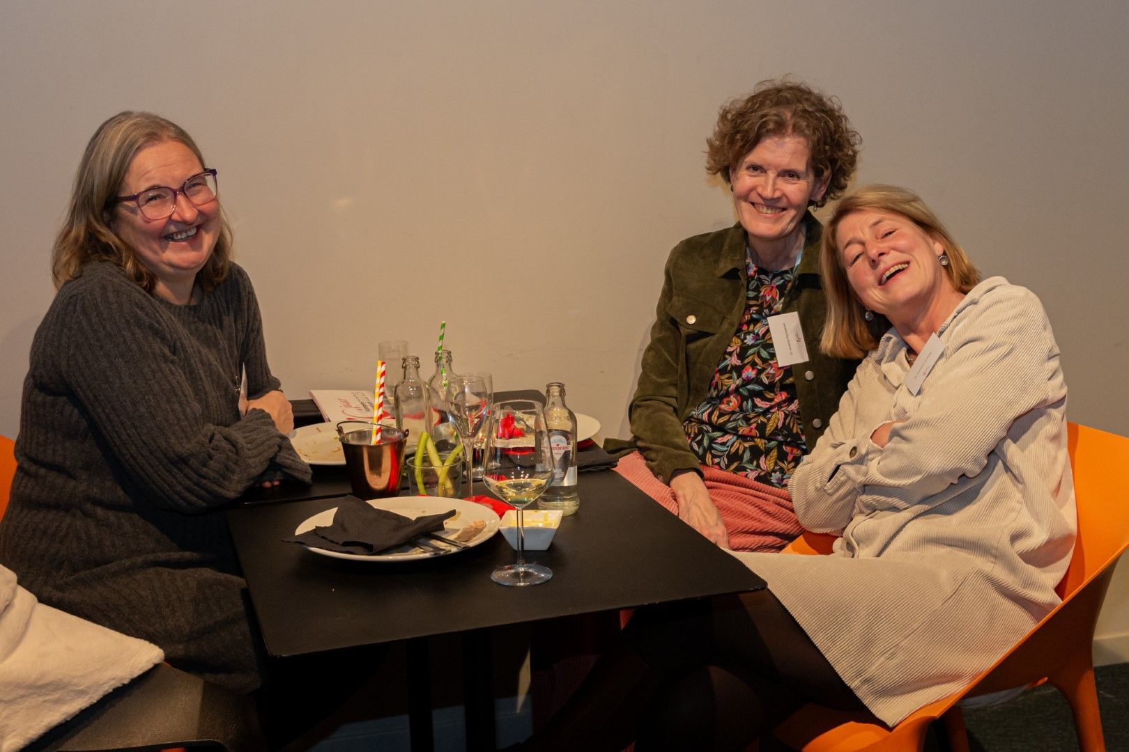 Drie mensen glimlachen aan een cafétafel met drankjes en borden, en poseren samen binnen.