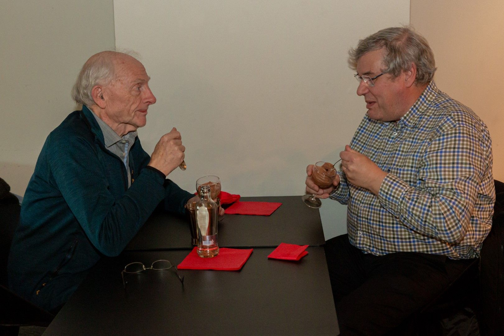 Twee mannen zitten aan een tafel en praten onder het genot van een drankje in een informele binnenomgeving.