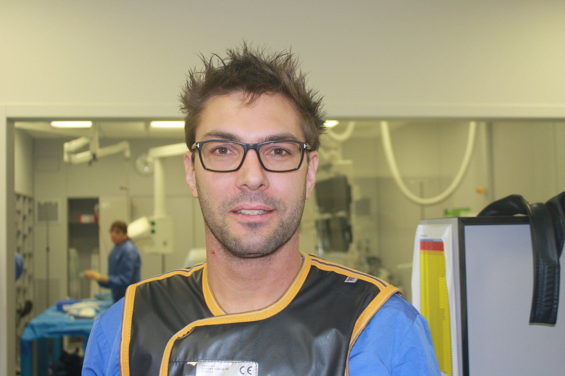 Een man met een bril en een zwart schort staat in een laboratoriumomgeving en kijkt in de camera.