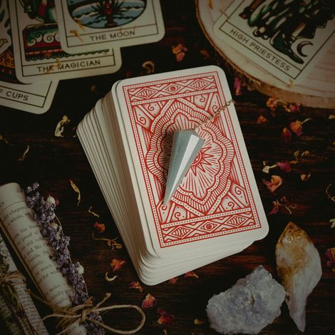 Tarot cards spread around a red-backed deck with a crystal pendulum on top and stones nearby