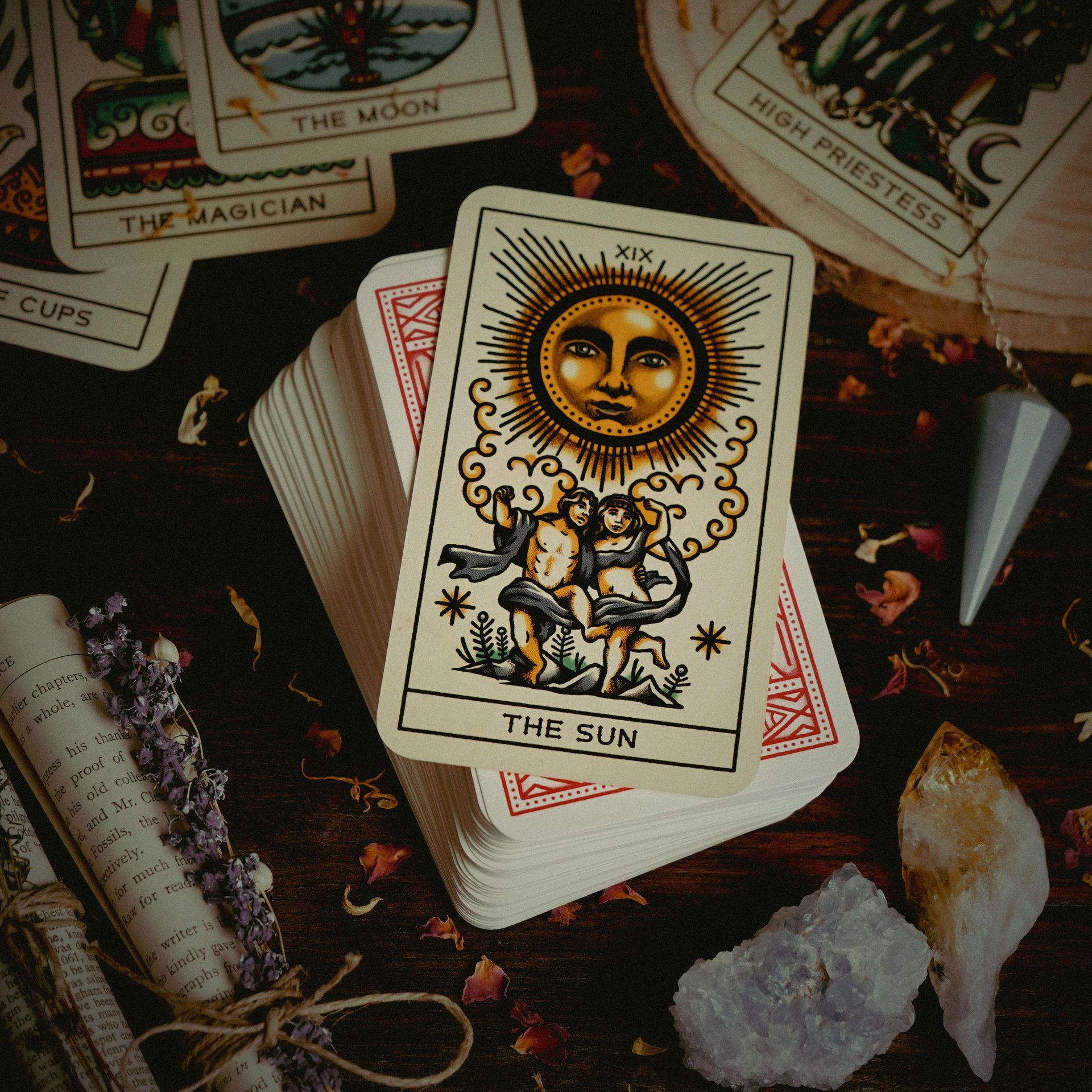 Tarot cards spread on a dark surface, with The Sun card centered among crystals and dried herbs.