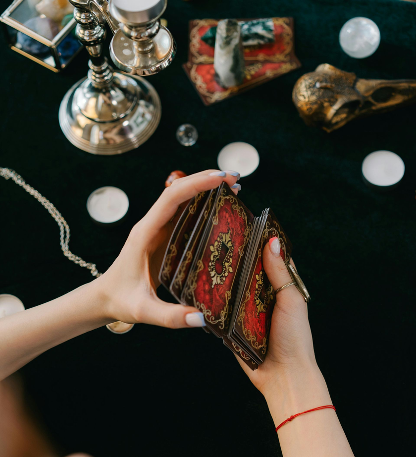 Hands holding a deck of ornate cards over a dark table with crystal balls and ritual objects