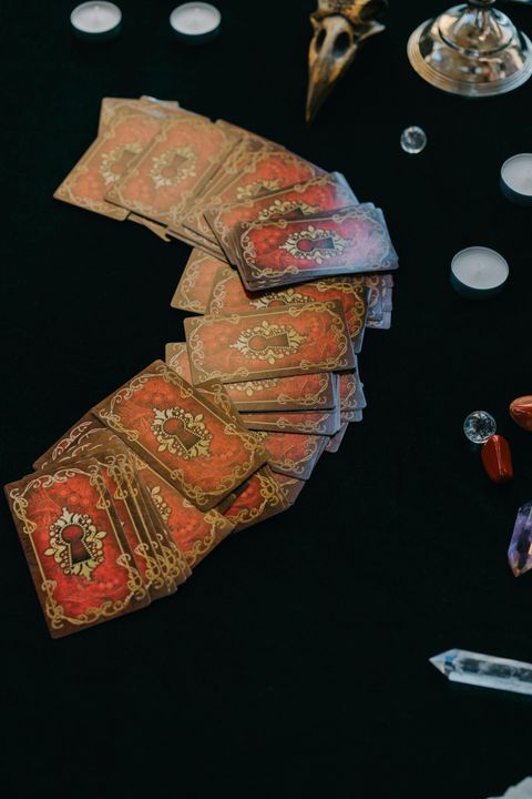 Fan of ornate red-orange playing cards on a dark surface with small white and glass objects nearby