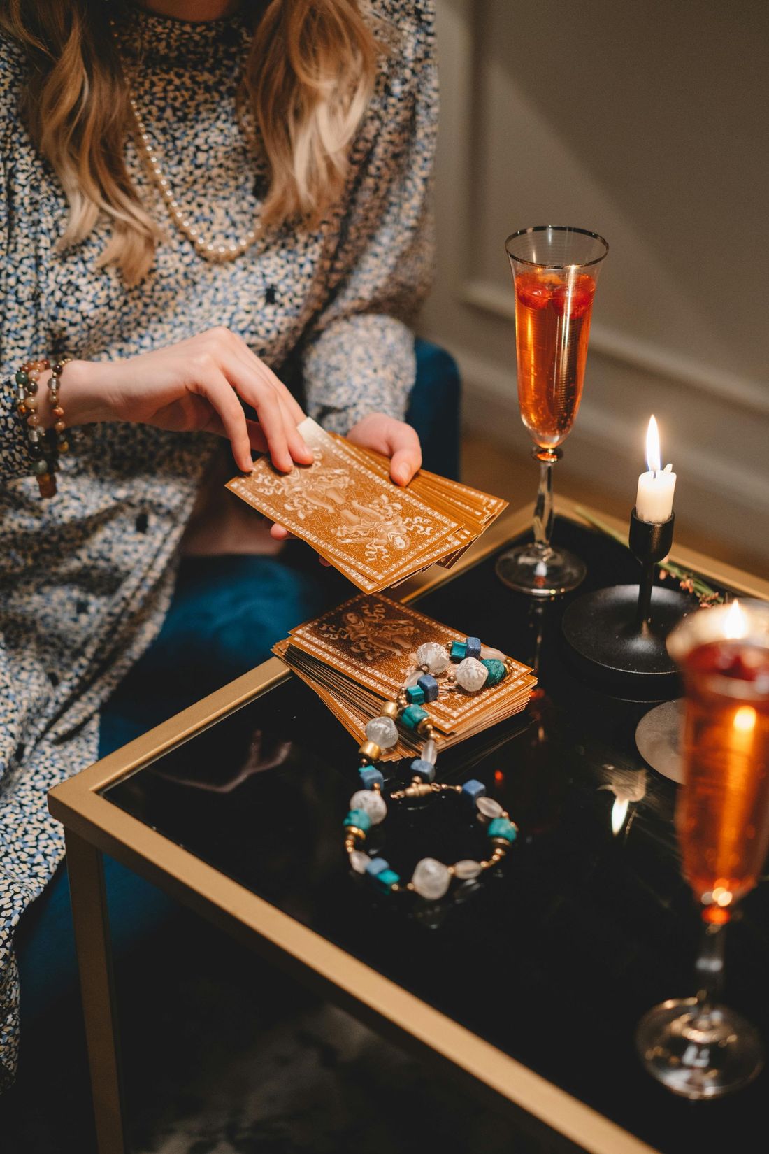 Person reading tarot cards at a candlelit table with jewelry and a glass of amber drink
