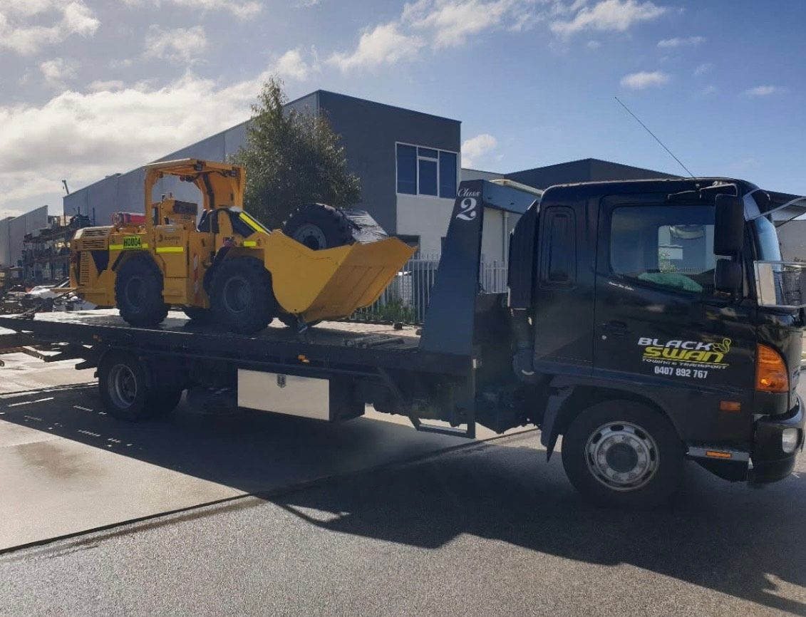 Yellow construction loader on a black flatbed truck, parked on a paved road. Blue sky.