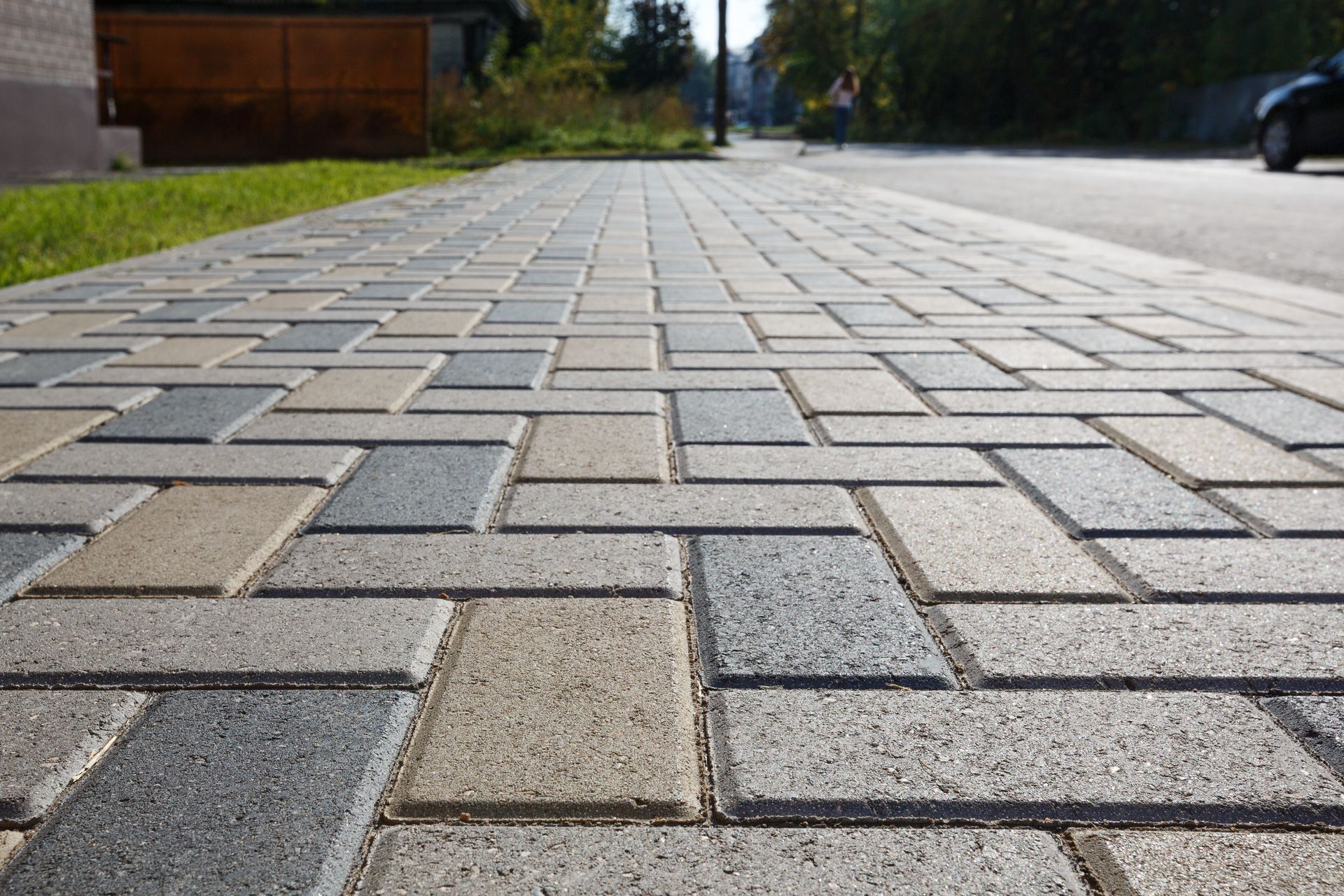 Low-angle view of a patterned brick sidewalk beside a road with grass and trees in the distance