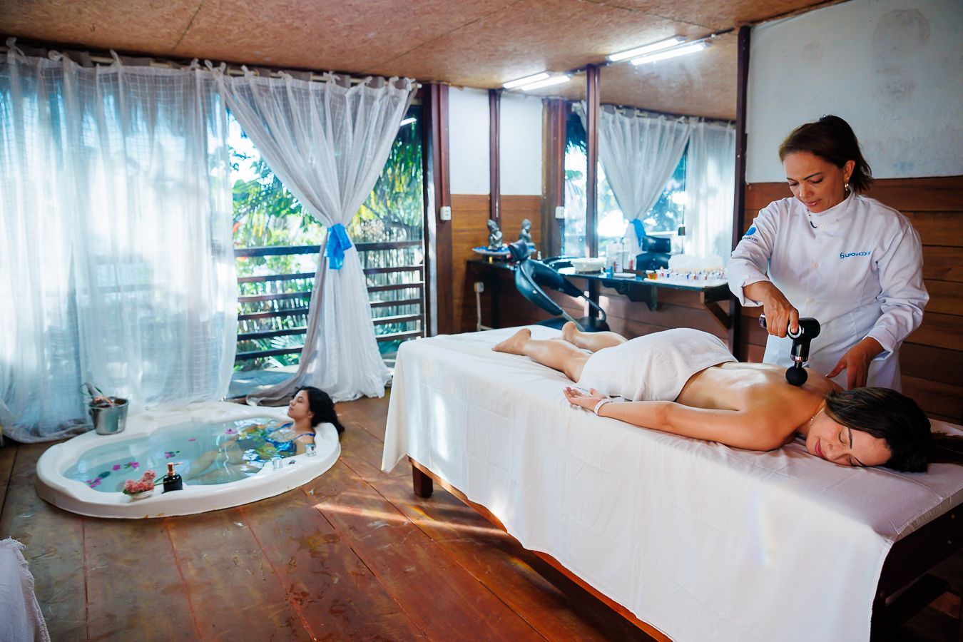 Sala de tratamento de spa com mesa de massagem, terapeuta e uma pequena banheira de hidromassagem.
