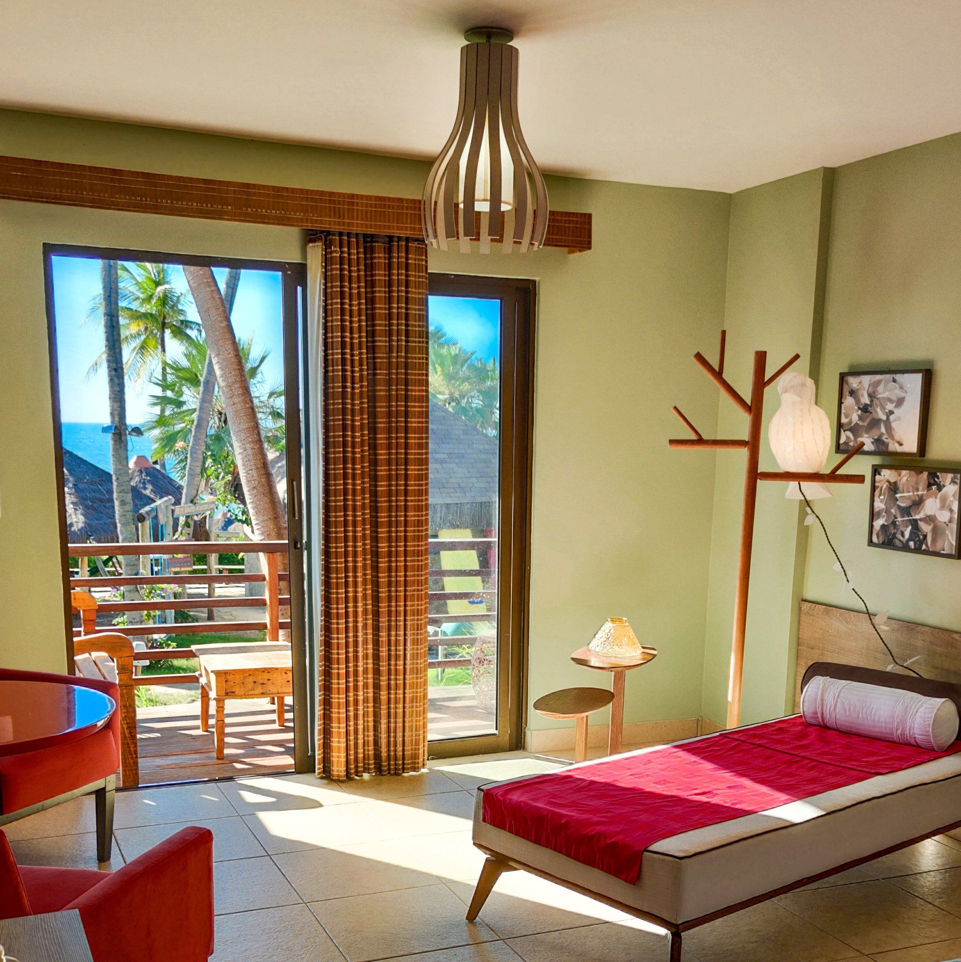 Quarto ensolarado com colcha vermelha, decoração em madeira e varanda aberta com vista para palmeiras.