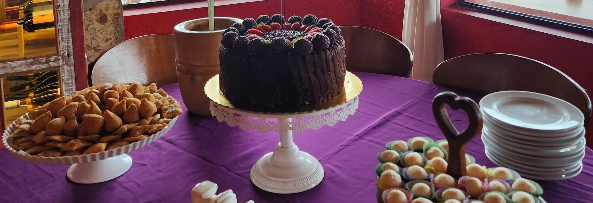 Mesa de sobremesas com bolo de chocolate, doces e pratos empilhados sobre uma toalha de mesa roxa.