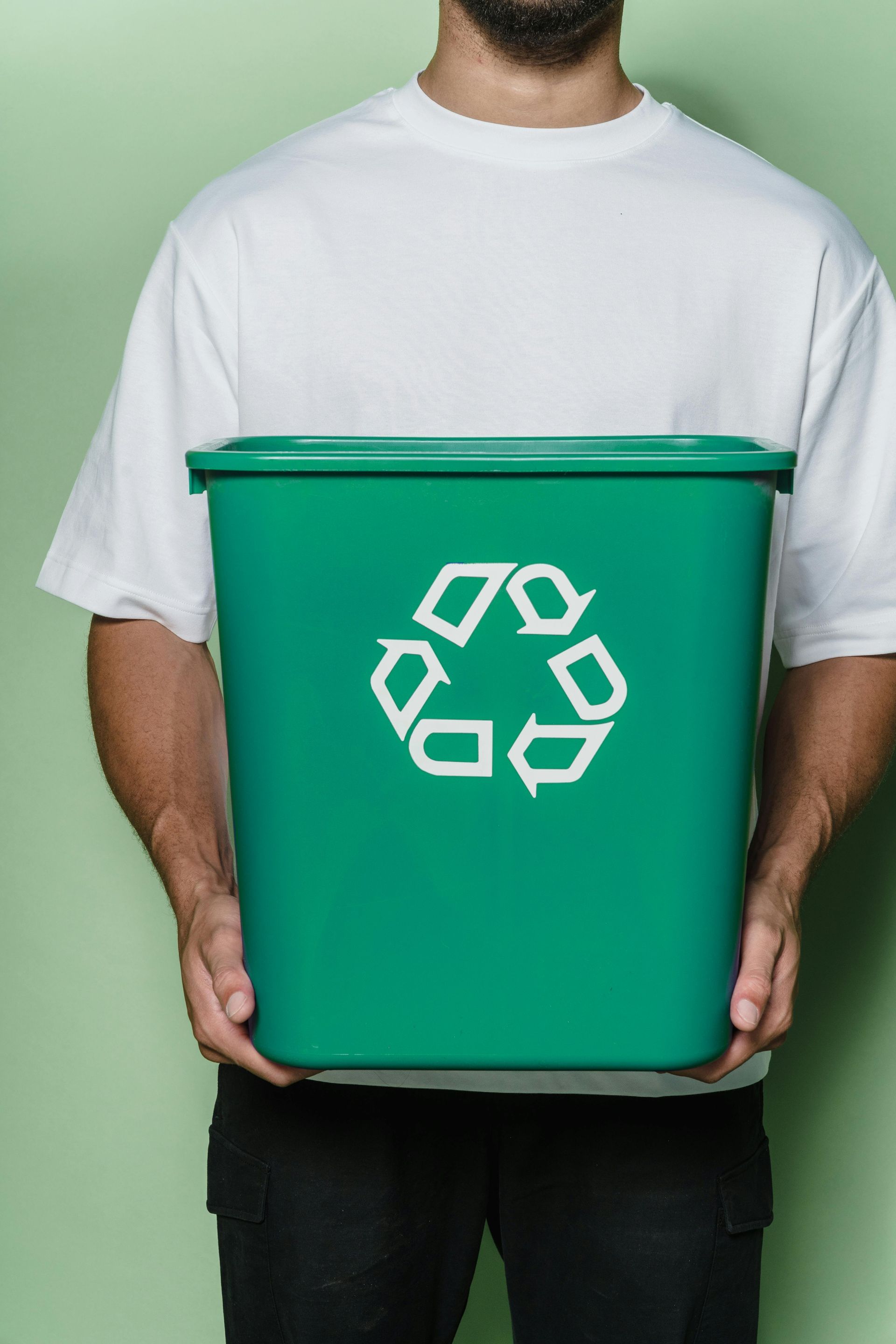 Pessoa segurando uma lixeira de reciclagem verde com um símbolo de reciclagem branco contra um fundo liso.