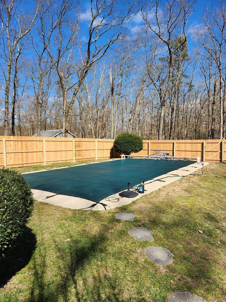 Pool covered with dark green tarp in a fenced backyard, surrounded by trees under a blue sky. Pool covered with dark green tarp in a fenced backyard, surrounded by trees under a blue sky.