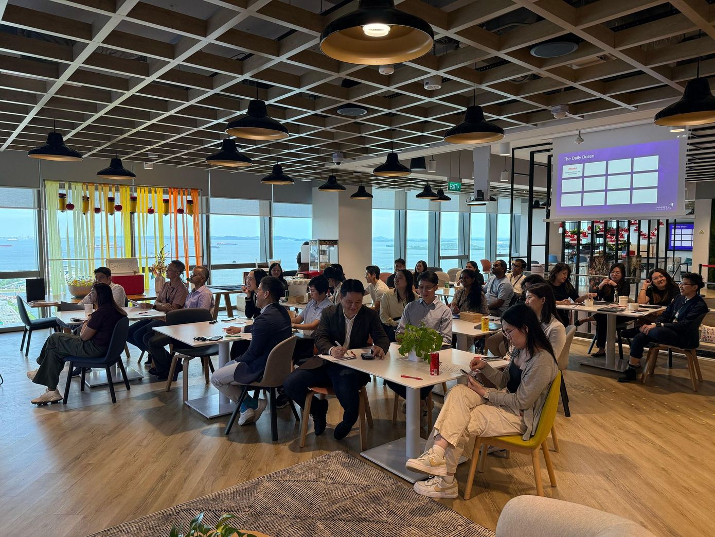 People seated at tables in a bright room with a presentation screen. They are likely attending a meeting or event.