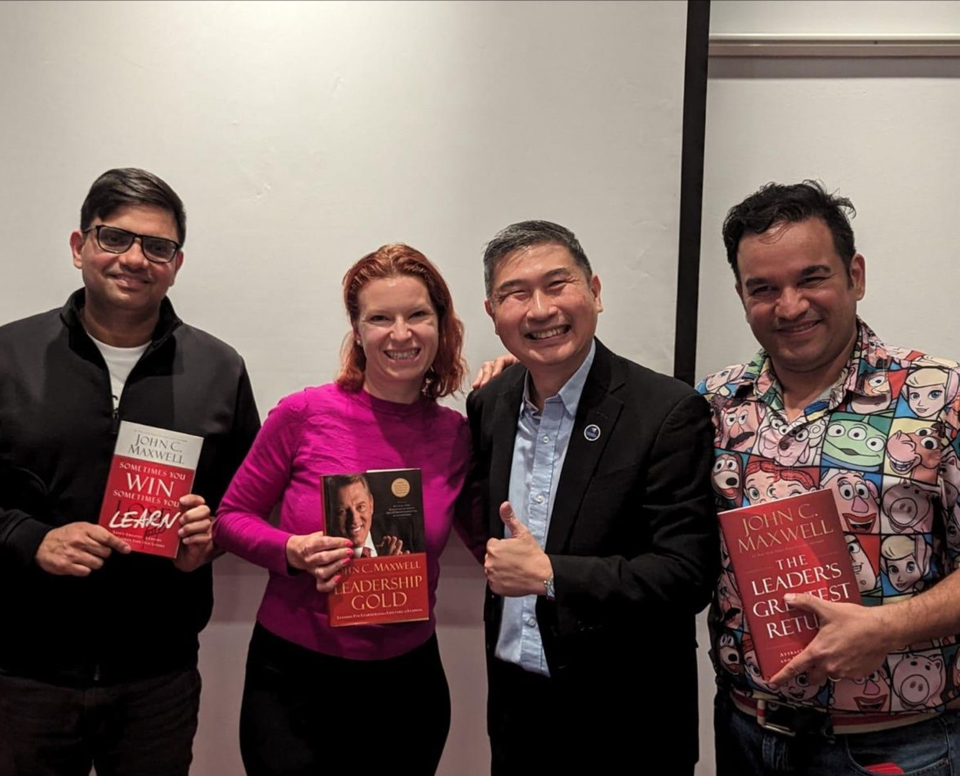 Four people pose with books; one man gives a thumbs-up. The setting is indoors with a white wall.