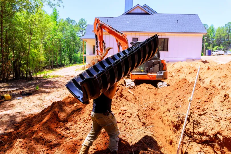 Excavator lifting a large black pipe into a trench beside a white house under construction