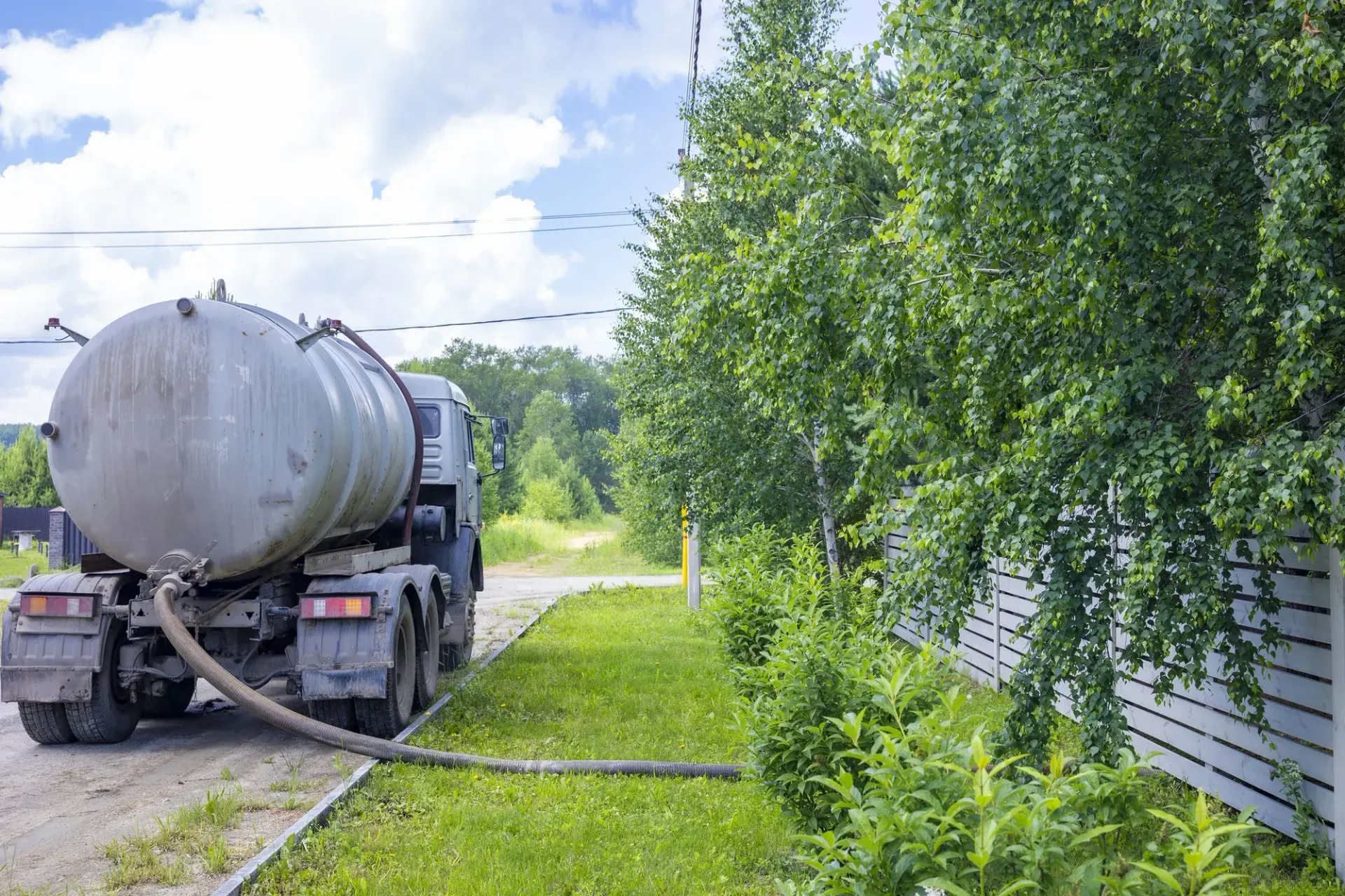 Septic tank truck parked on a grassy roadside beside trees and a house, pumping waste through a hose