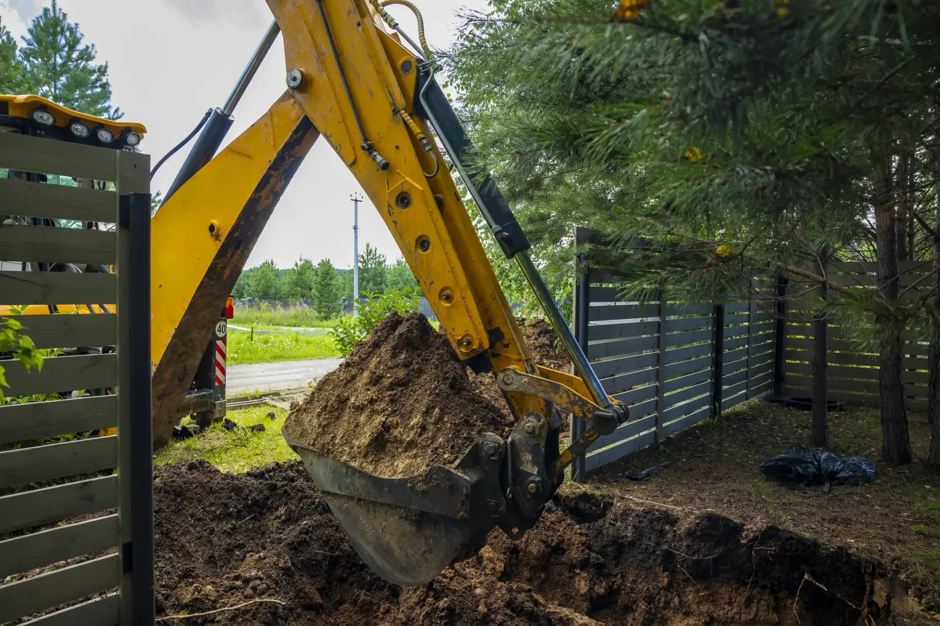 Excavator digging soil beside a fence in a muddy yard with trees nearby