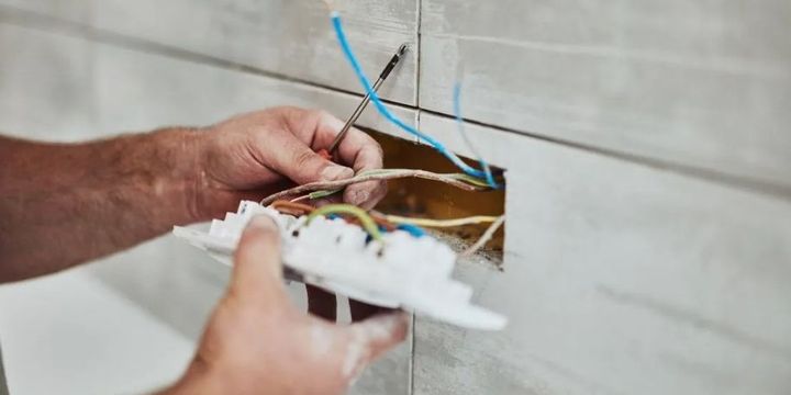 Hands wiring a white electrical connector with blue, brown, and yellow cables against a wall panel