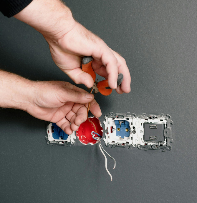 Hands wiring an exposed electrical outlet and switch on a gray wall, with red and blue junction boxes.