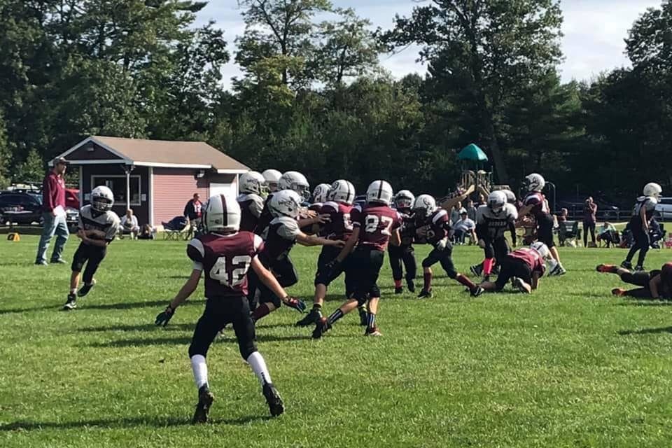 Youth football game on a grassy field, players in black and white uniforms near a small sideline building.