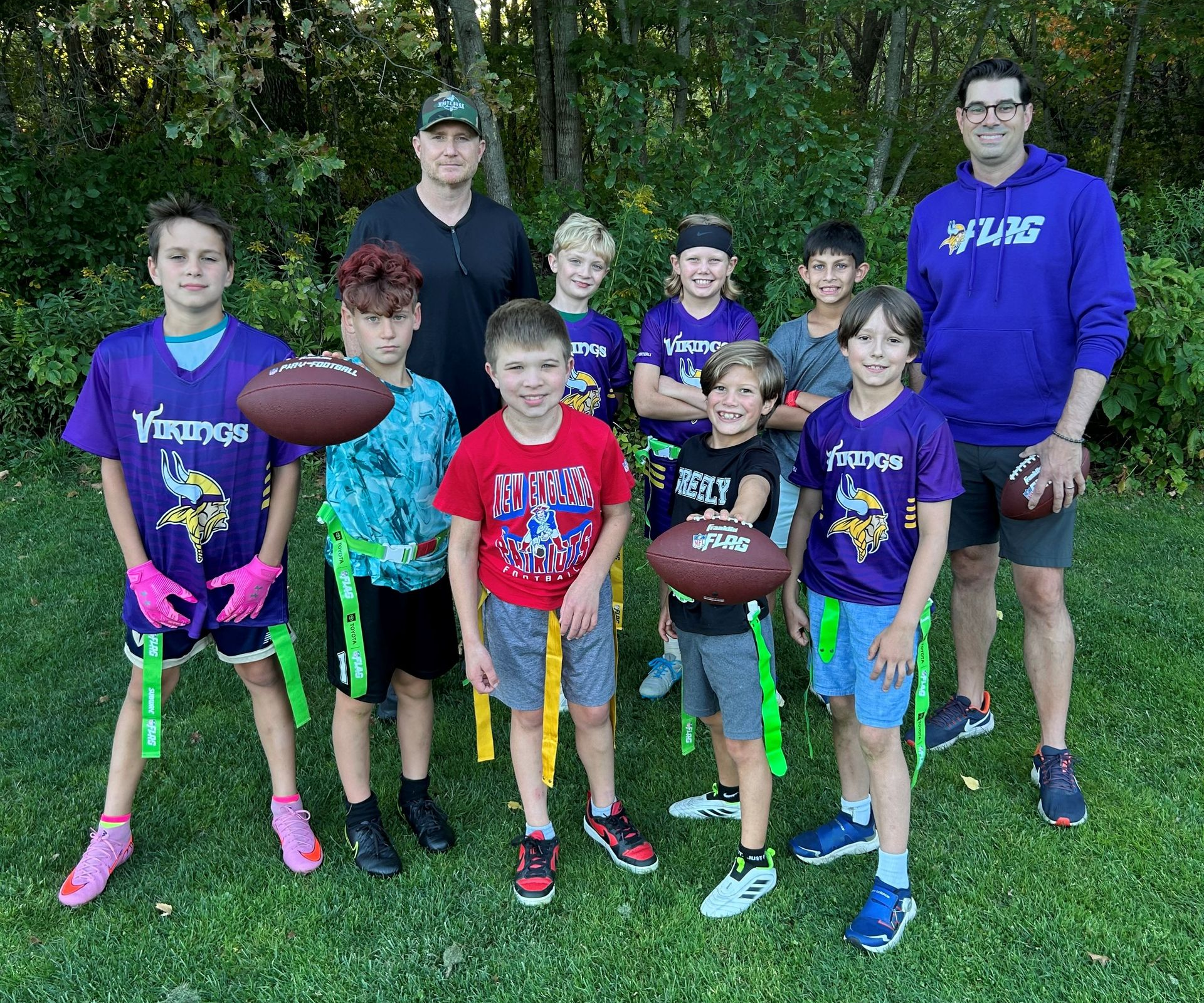 Youth football team and coaches posing on grass, holding footballs, in purple jerseys with trees behind them