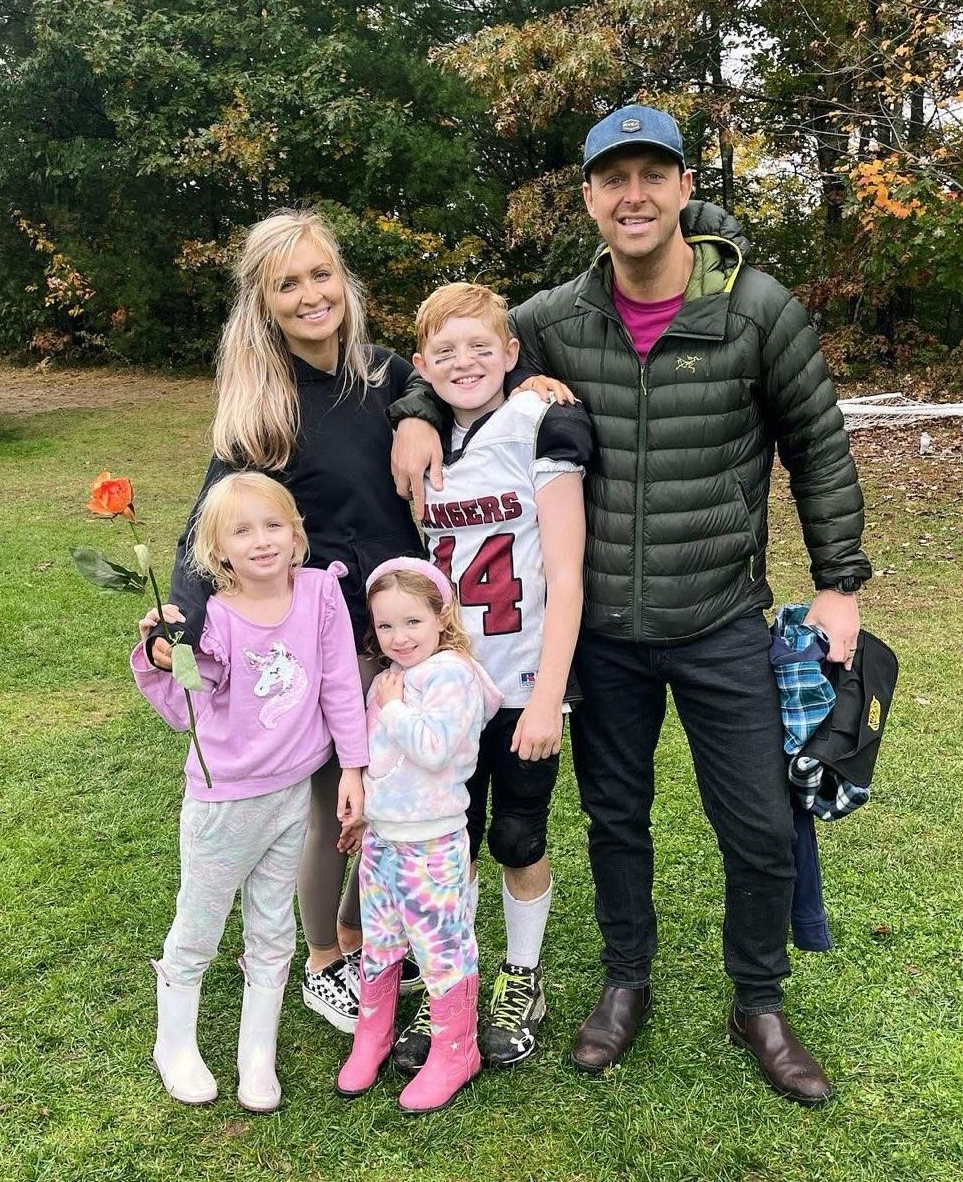 Family of four outdoors on grass, smiling and standing together in autumn with trees behind them.