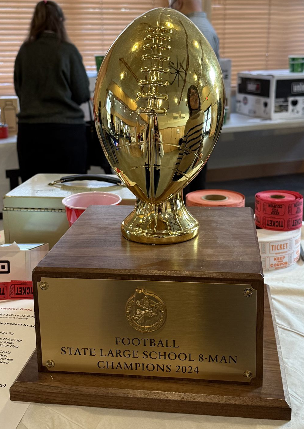 Gold football trophy on a wooden base on a table, with a person and office items blurred in the background