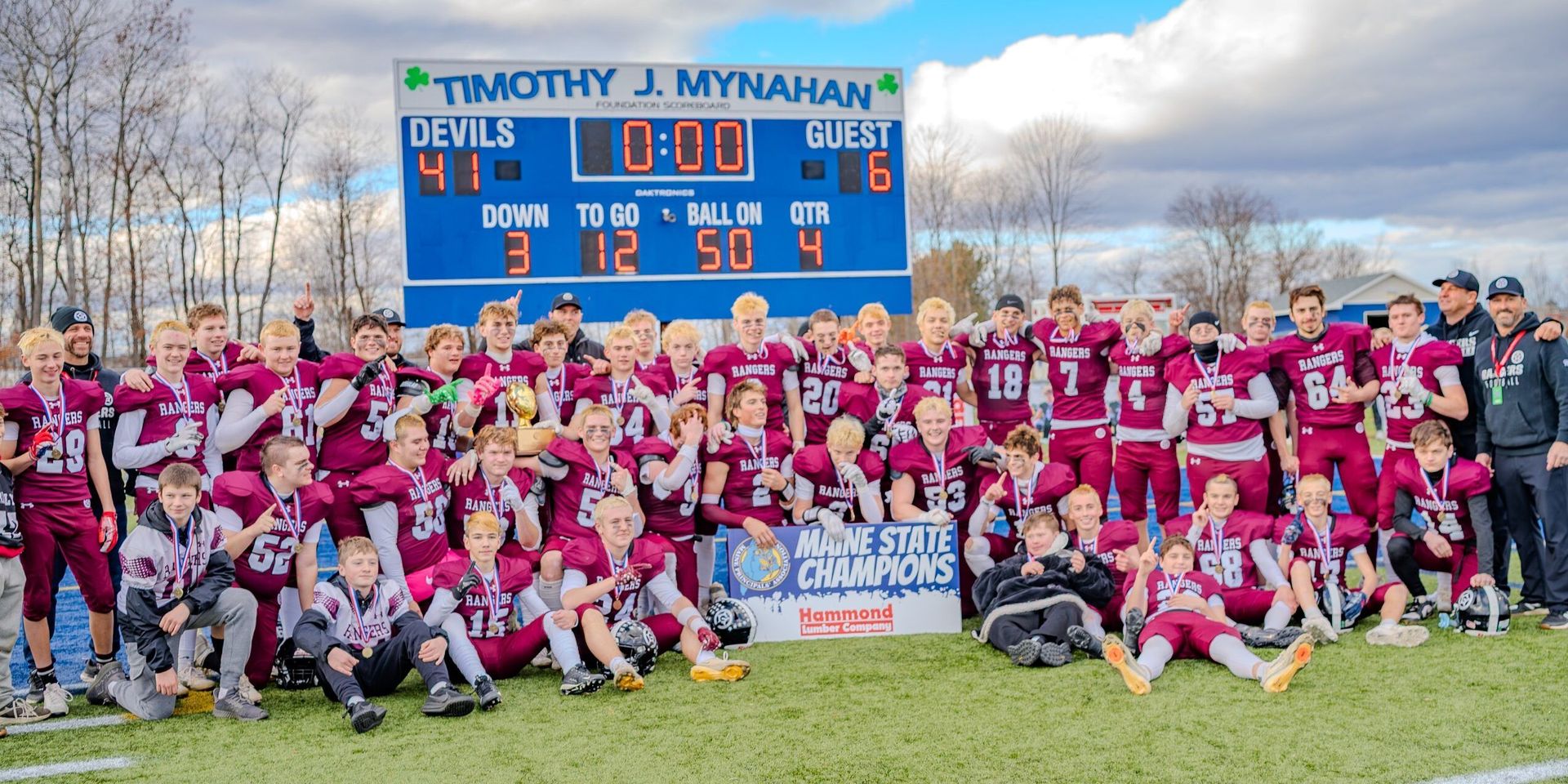 Youth sports team in maroon uniforms posing on a football field before a scoreboard sign