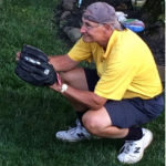 A man in a yellow shirt is squatting down in the grass holding a baseball glove.