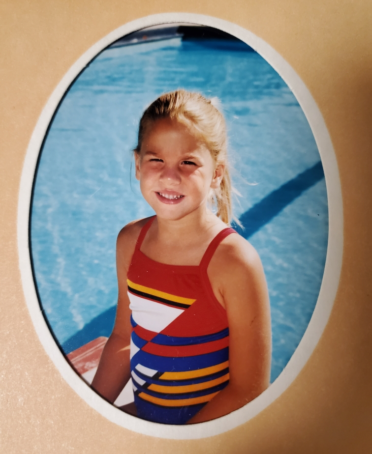 A young girl in a red and blue striped swimsuit sits in front of a swimming pool
