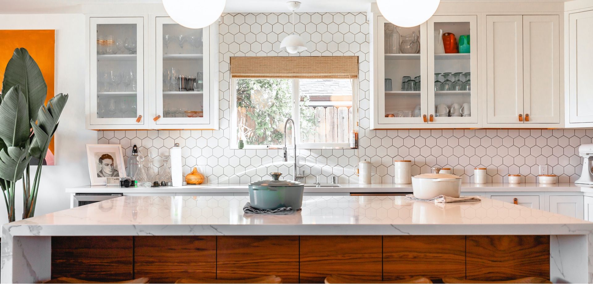 Modern kitchen with white cabinets, marble countertop, and wood island. Green plant and orange artwork.